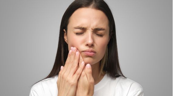 Young woman with long dark hair wearing a white shirt, holding her cheek with a pained expression, indicating a possible toothache or dental pain